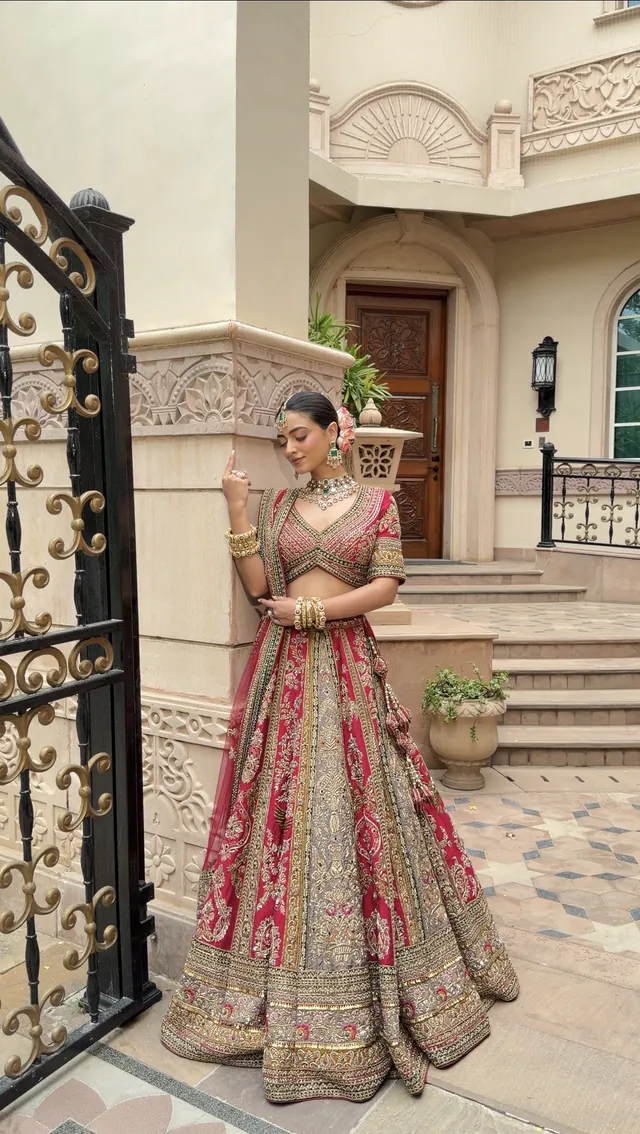 Bride in a red and gold embroidered lehenga, pausing beside an ornate iron gate