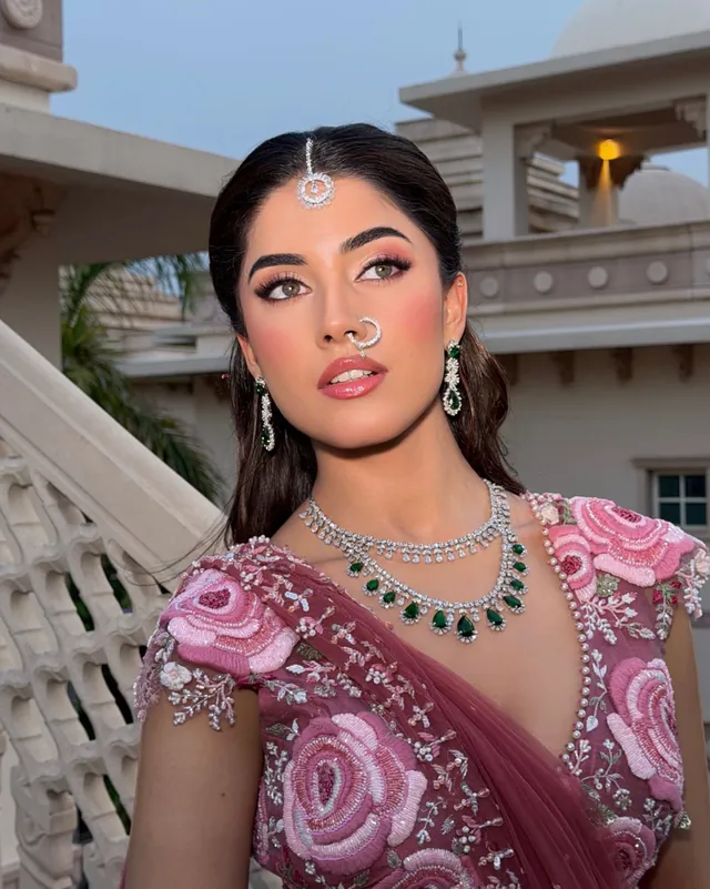 Bride in a rose pink floral-embroidered lehenga with emerald and diamond jewellery, in a palace courtyard at dusk